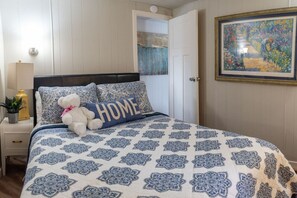 Un dormitorio con una cama que tiene una colcha azul y blanca estampada, un oso de peluche blanco y una almohada con la palabra 'HOME' escrita.
