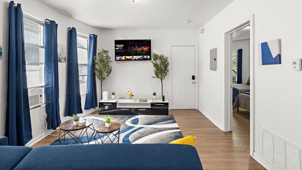 Un salón moderno con un sofá, una mesa de centro y un televisor montado en la pared. Se observa una puerta que da a otra habitación y una ventana con cortinas.