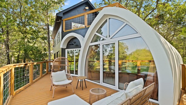 Une maison moderne dotée d’une terrasse en bois, de meubles en osier blancs et de grandes fenêtres en verre.