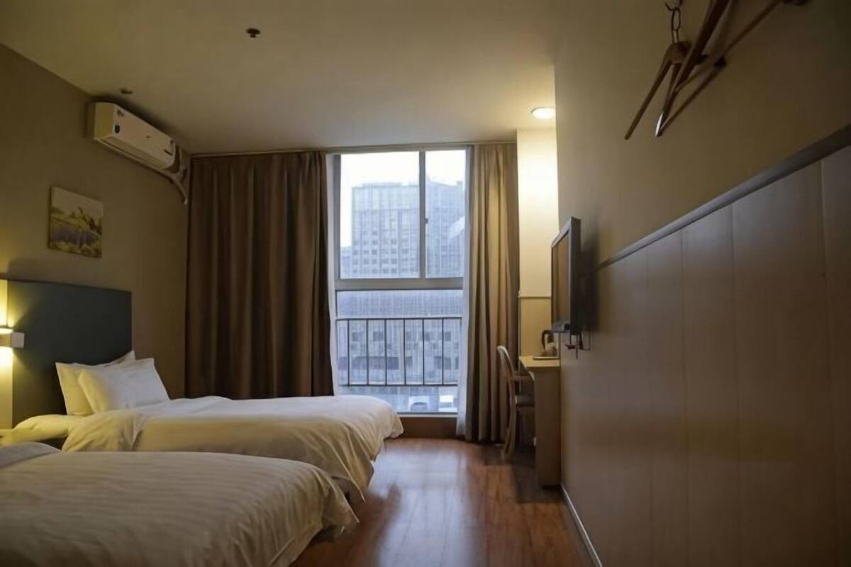 A hotel room with two beds, a desk, a chair, a TV, and a window with a view of buildings.