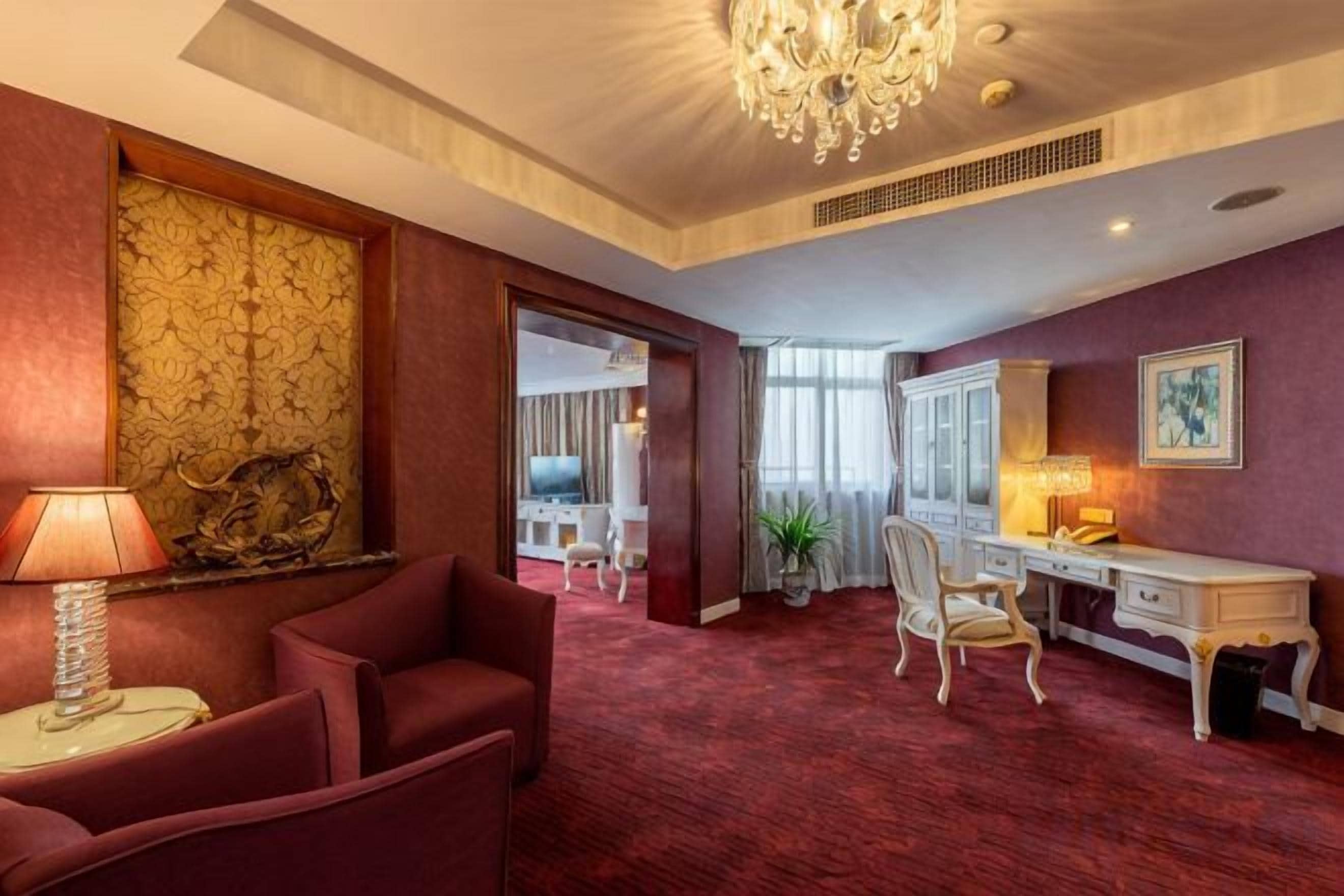 A hotel room with a red carpet, a desk with a lamp, a chair, a chandelier, and a large window.
