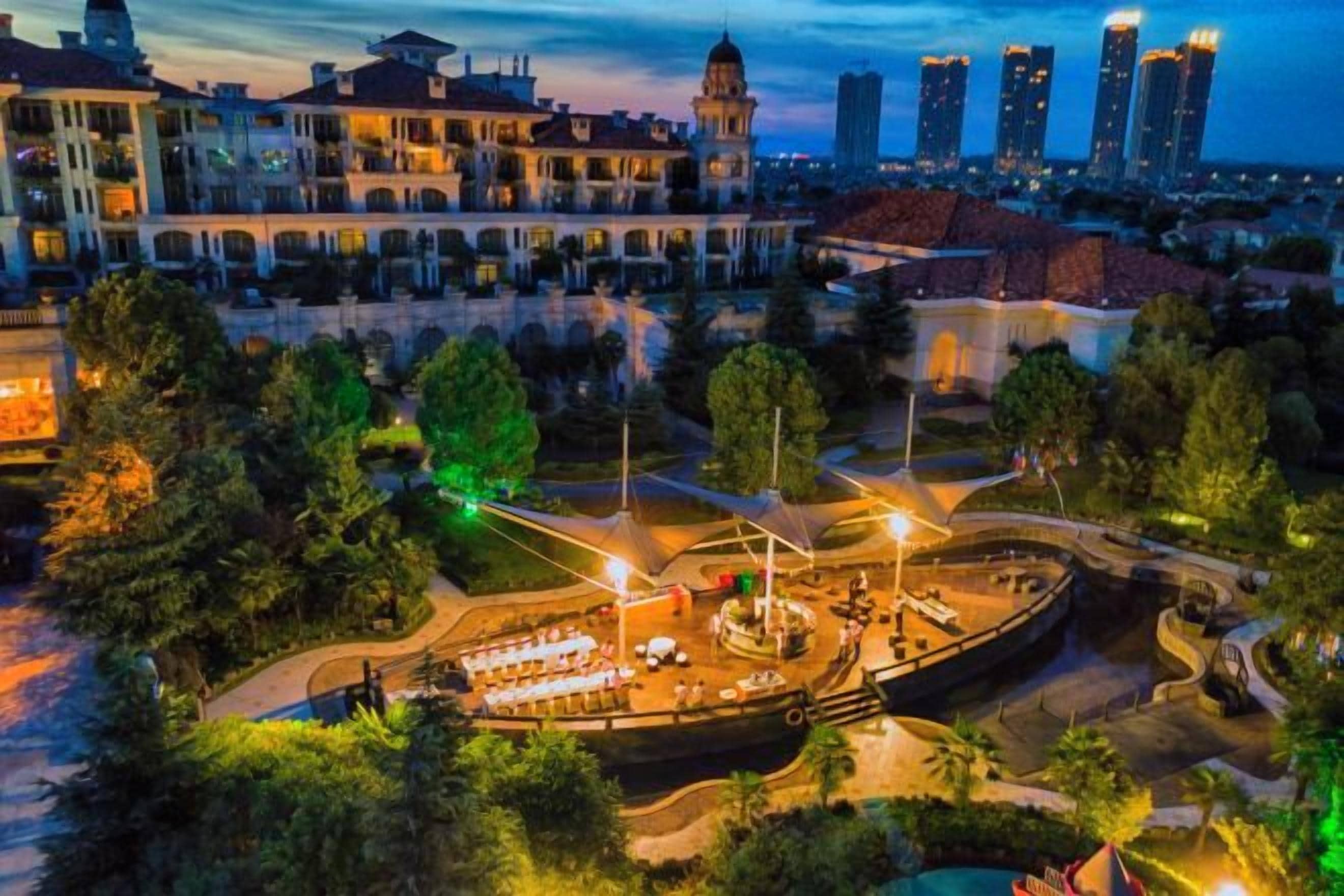An aerial view of a historic building complex with a central courtyard, surrounded by trees and modern high-rise buildings in the background.
