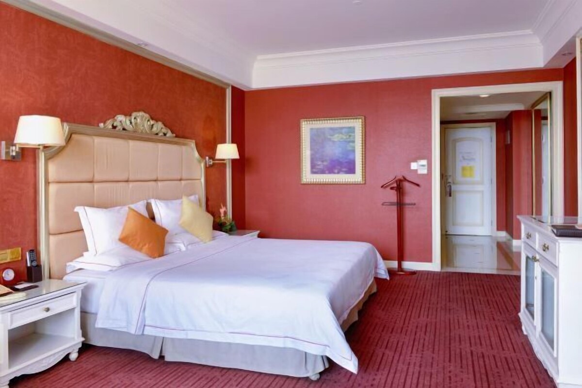 A hotel room with a large bed, a nightstand, a lamp, a painting on the wall, and a door leading to another room.