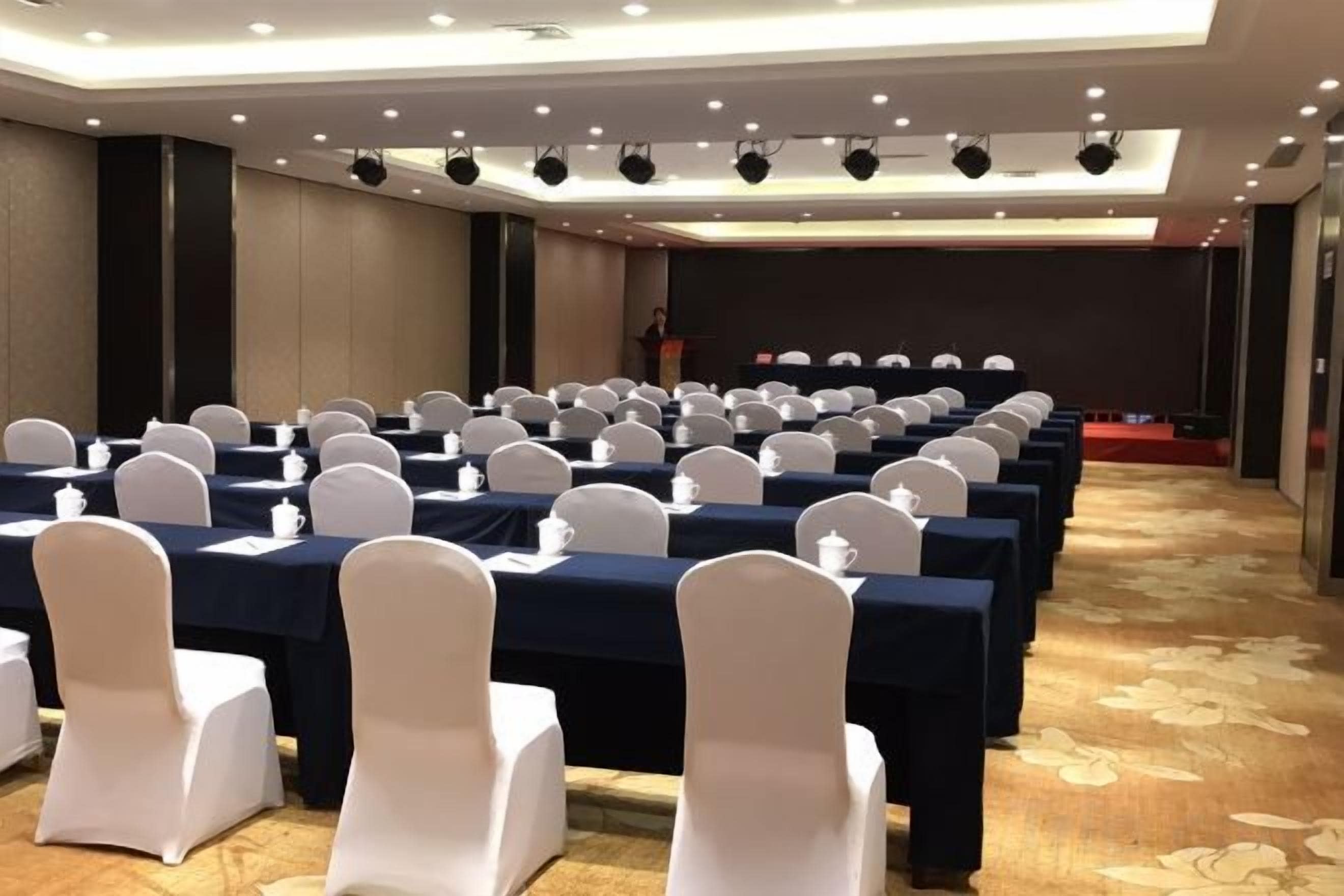 A conference room with rows of tables and chairs, a stage, and a carpeted floor.