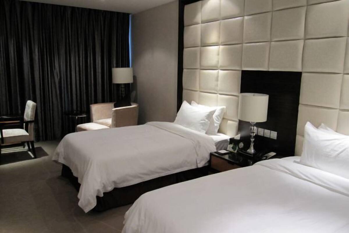 A hotel room with two beds, a large padded headboard, a desk with a chair, and a lamp.