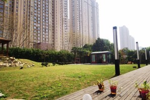 Area verde con una veranda in legno, piante in vaso e un edificio sullo sfondo.