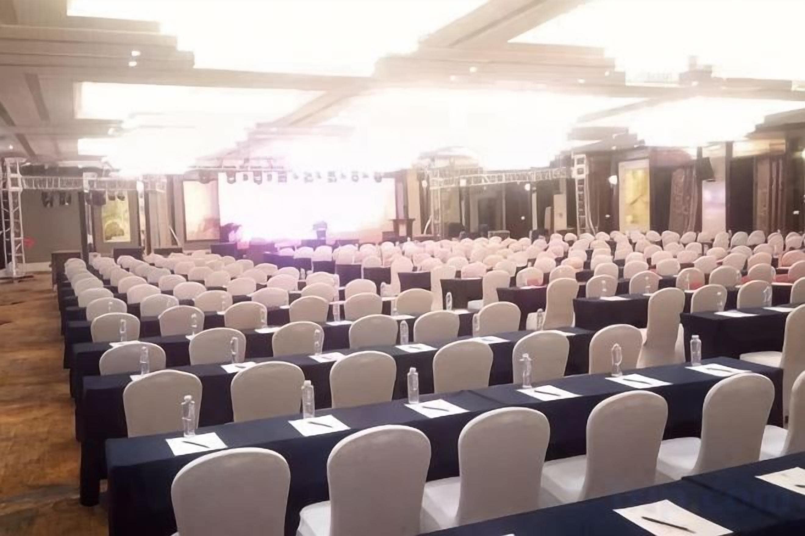 A conference room with rows of tables and chairs, a stage with a screen, and a large window.