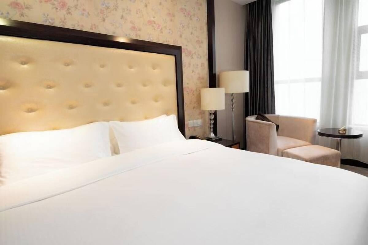 A hotel room with a large bed, a headboard, a bedside table with a lamp, a chair, and a small table.