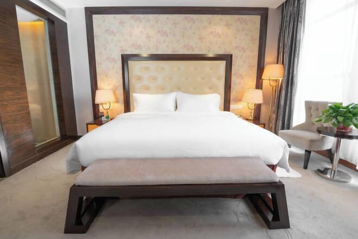 A hotel room with a large bed, two bedside lamps, a bench at the foot of the bed, a chair, and a small table with a plant.