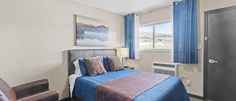 A hotel room with a bed, a chair, a painting, a window with curtains, and a door.