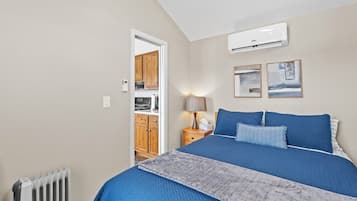 A bedroom with a large bed, a radiator, a nightstand with a lamp, and a door leading to a kitchen.