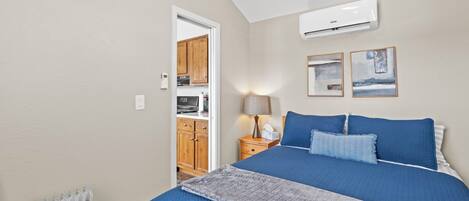A bedroom with a large bed, a radiator, a nightstand with a lamp, and a door leading to a kitchen.