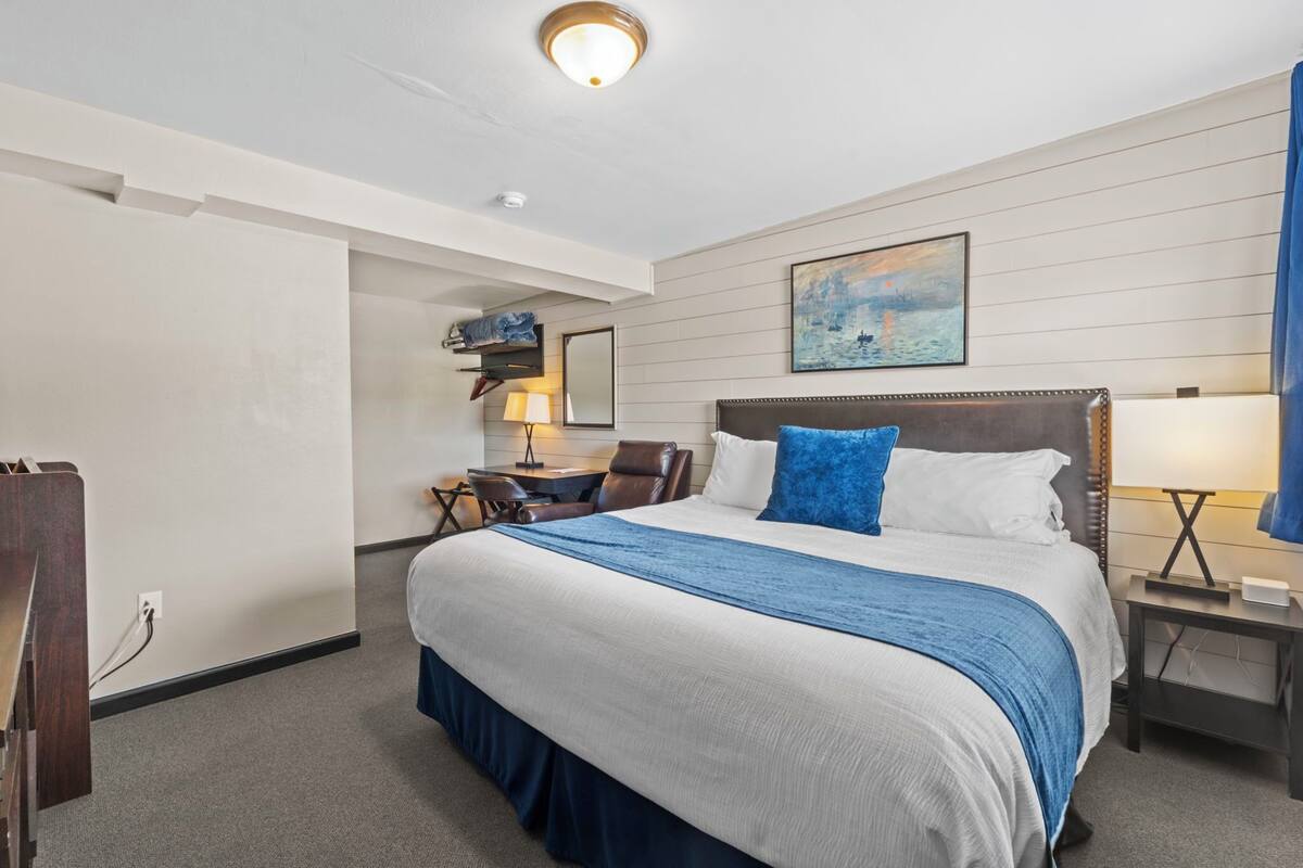 Ein Hotelzimmer mit einem großen Bett, einem Schreibtisch mit Stuhl, einer Schreibtischlampe, einem Nachttisch und einem Bild an der Wand.