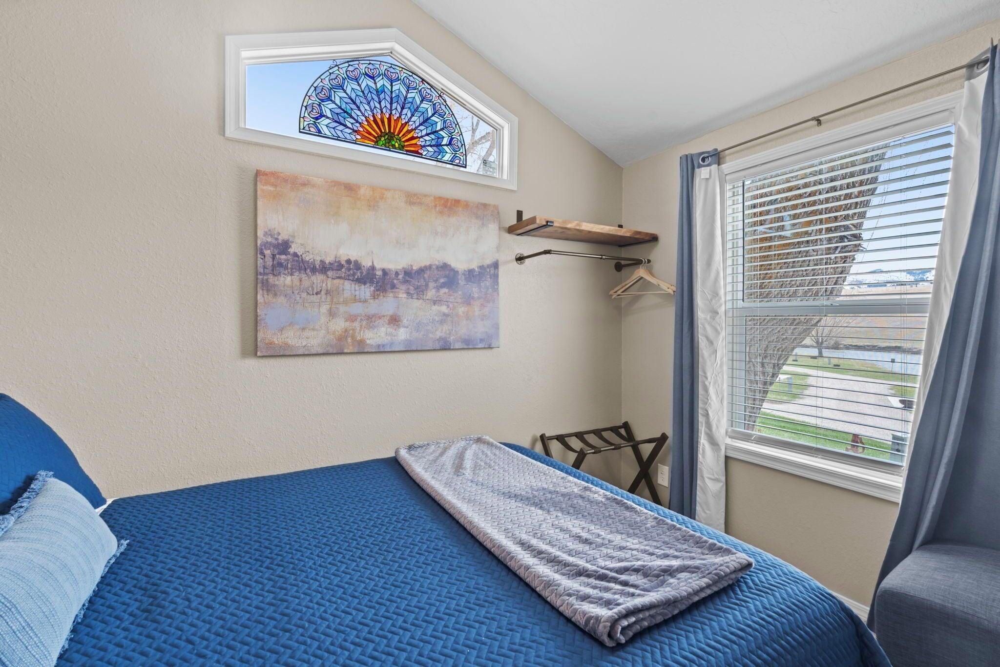 A bedroom with a bed, a window with blinds, a wall-mounted artwork, and a ceiling light fixture.