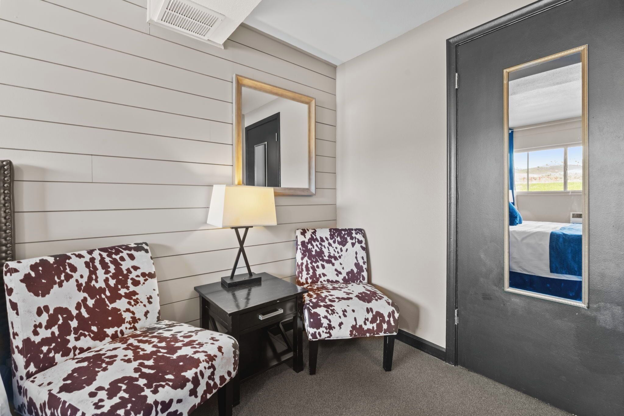 A hotel room with a bed, a desk with a lamp, two patterned chairs, a mirror, and a door with a window view.