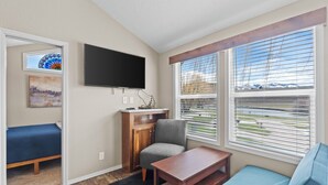 A compact living space with a bed, a TV mounted on the wall, a small table, and a window with blinds. - Lost Trail Resort (Lava Hot Springs)