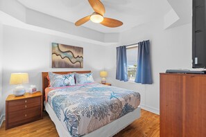 A bedroom with a bed, wooden nightstands, a ceiling fan, a painting, a window with curtains, and a wardrobe.