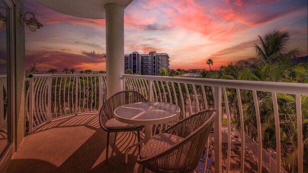 A balcony with wicker chairs and a small table, overlooking a cityscape with buildings and palm trees under a colorful sunset sky. - Beach Gear Included! Crisp, Clean & Great Value (Treasure Island)