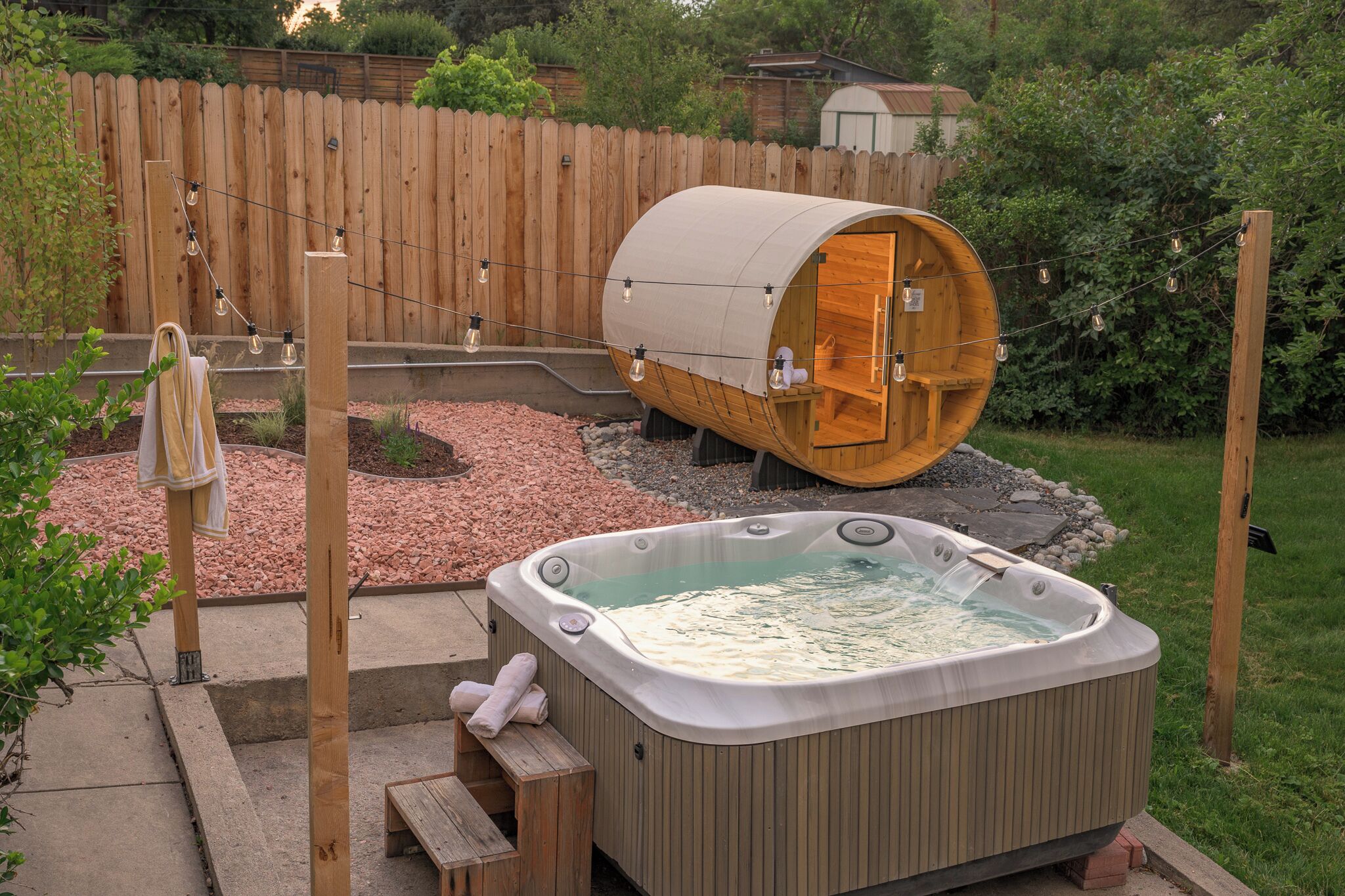 Un bain à remous et un tonneau en bois dans un jardin, avec une clôture en bois et de la verdure.