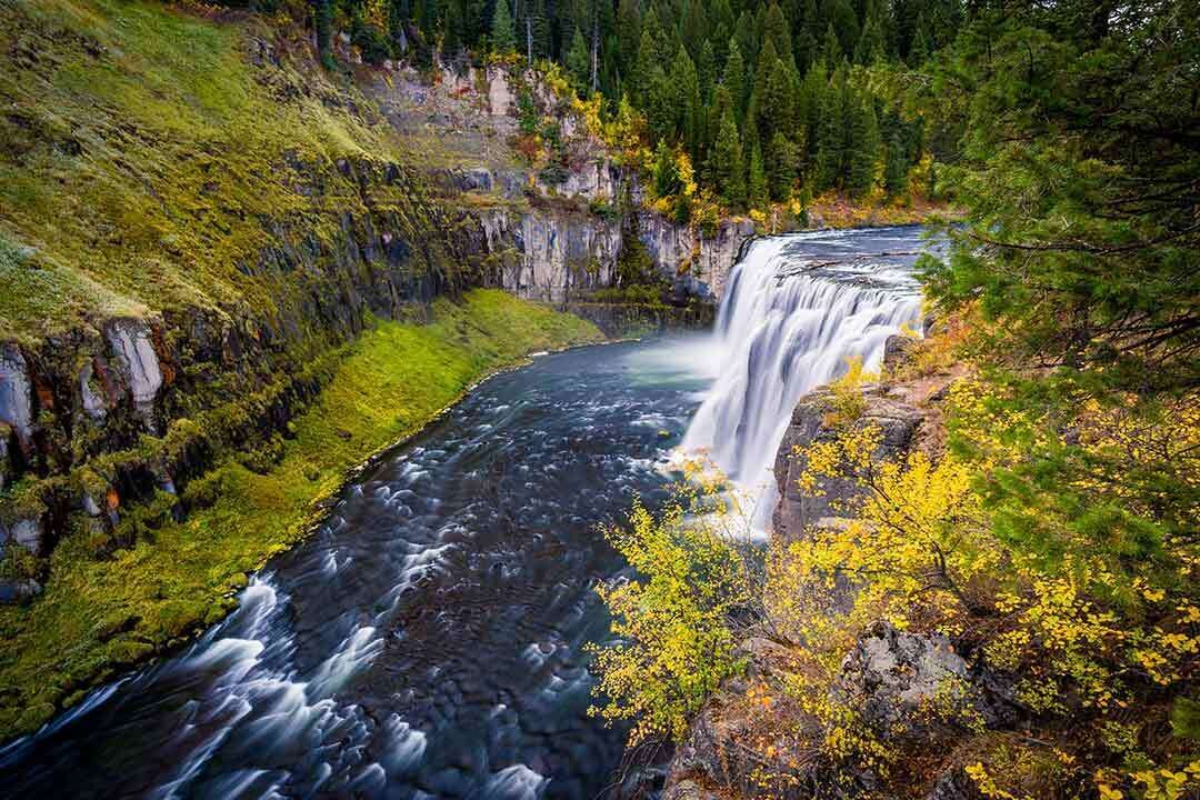 A waterfall cascading into a river surrounded by lush greenery and autumn foliage.