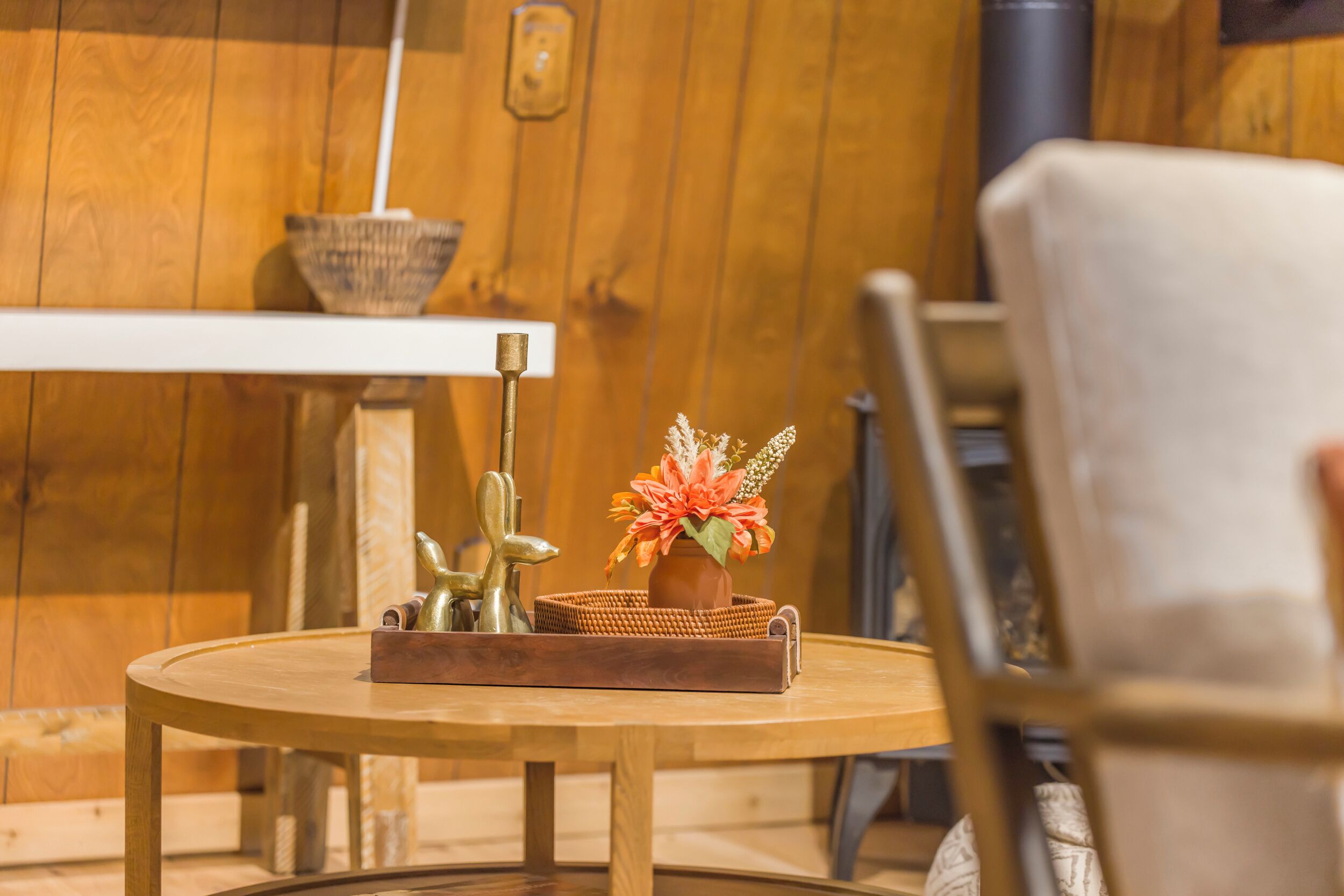 Een ronde houten tafel met een gevlochten dienblad waarop een terracotta pot met oranje bloemen en een messing sculptuur staan. Een witte stoel is gedeeltelijk in de voorgrond zichtbaar.