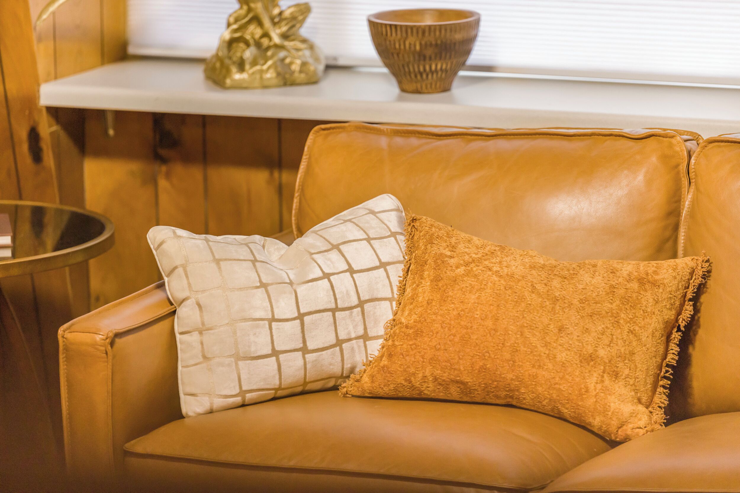 A tan leather sofa with two cushions, a small round table, and a decorative bowl on a white shelf.