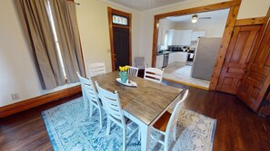 Un comedor con mesa de madera y sillas blancas, una cocina al fondo y una puerta con vitral.