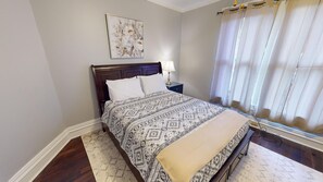 A bedroom with a bed, a nightstand, a lamp, a window with curtains, and a painting on the wall.