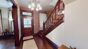 Una escalera de madera con intrincados grabados, una puerta de madera con un vitral, y una araña con múltiples bombillas.