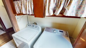 A compact laundry area with a front-loading washer and a top-loading dryer, both white, in a small room with wooden walls and a window with patterned curtains. - Historic Home in Nelsonville OH near Hocking hills (Nelsonville)