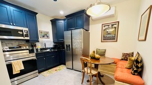 Una cocina compacta con armarios azul oscuro, un refrigerador de acero inoxidable, un microondas y un horno. Hay una mesa de comedor con sillas y una alfombra pequeña en el suelo.
