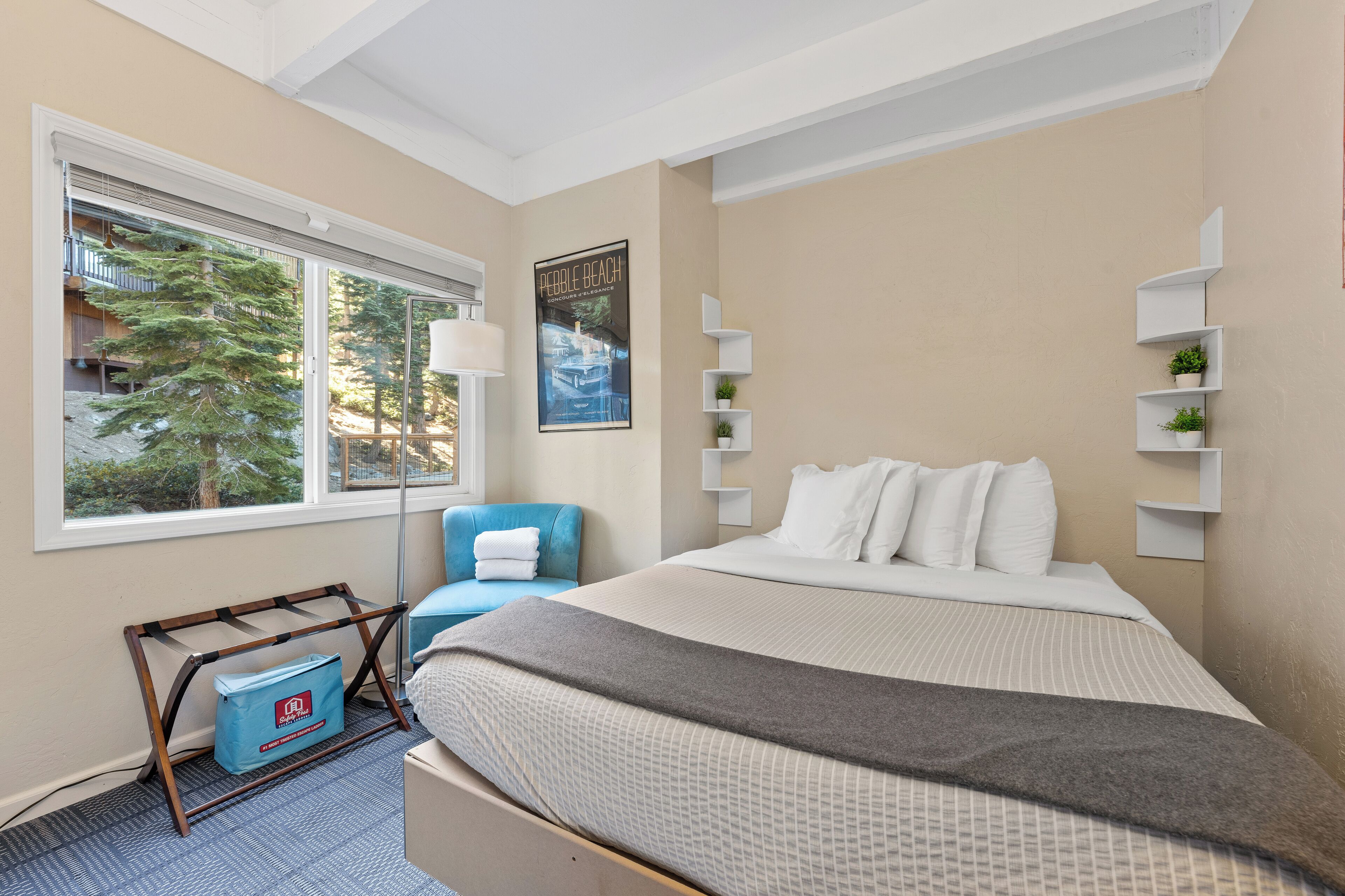 A bedroom with a bed, a window with a view of trees, a blue chair, and a wall-mounted shelf with plants.