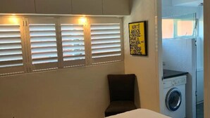 Un dormitori amb llit, una cadira, un ventilador de sostre, una finestra amb persianes i una rentadora a l'habitació veïna.
