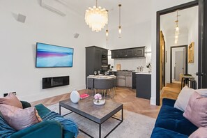 A modern living room with a sofa, coffee table, and a TV. There's a kitchen with dark cabinets and a dining area with chairs.