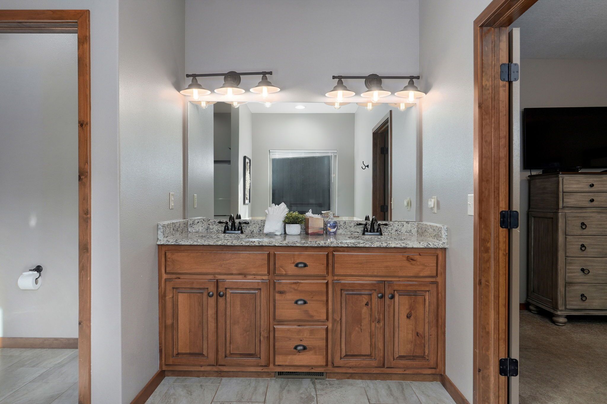 Une salle de bain dotée d’un meuble-lavabo double, d’armoires en bois, d’un grand miroir et d’un luminaire placé au-dessus du miroir.