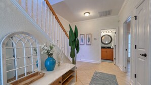A hallway with a wooden staircase, a white console table with a vase and mirror, a large plant, and a kitchen area with wooden cabinets and a round mirror. - Walk to the Beach with Pool and Game Room (Myrtle Beach)