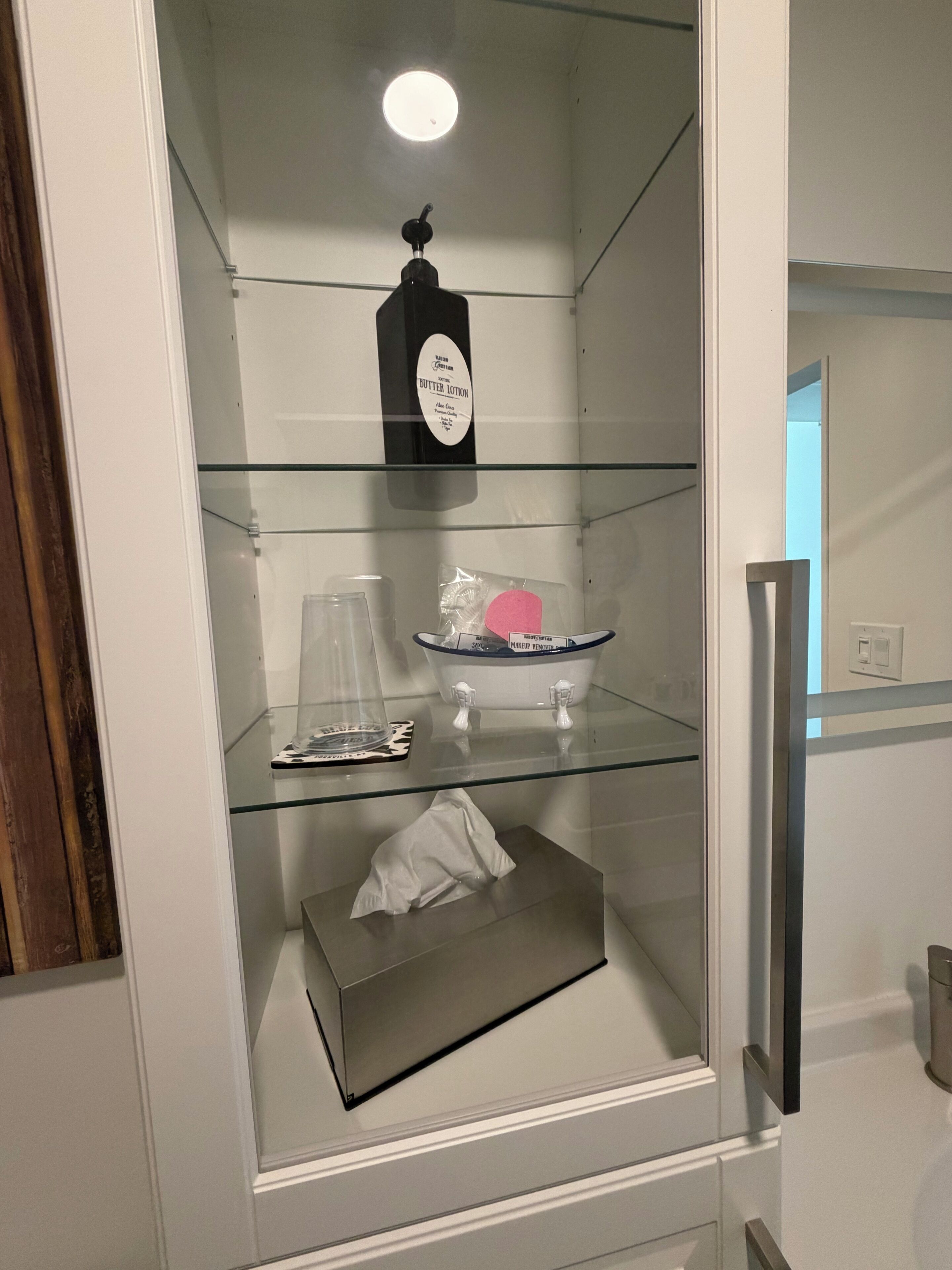 A bathroom with a glass shelf containing a bottle of lotion, a soap dish, and a tissue box.