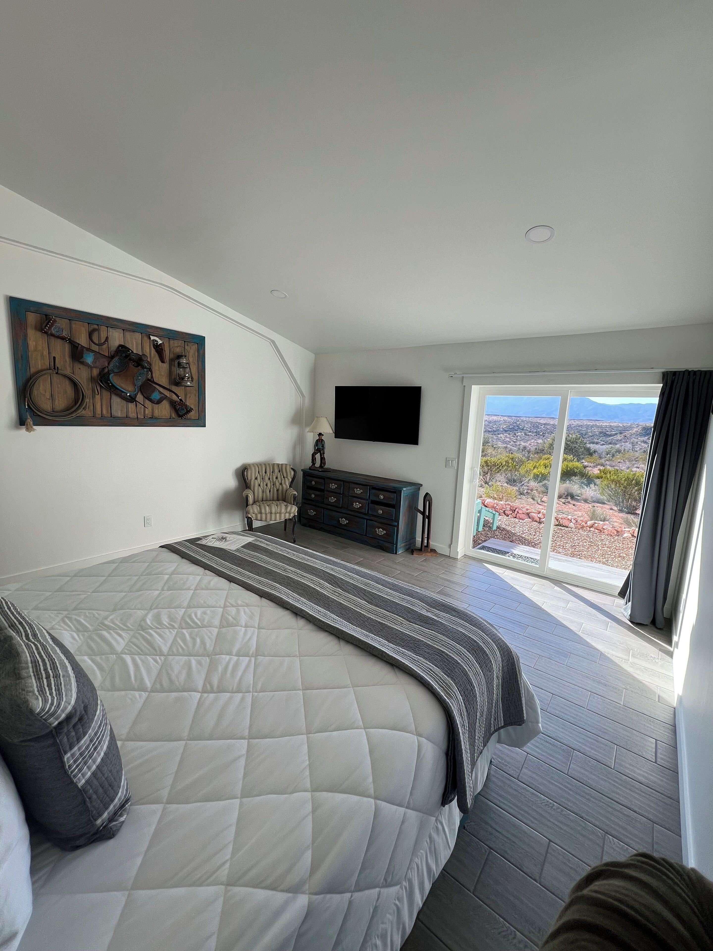 Un dormitorio con una cama grande, un aparador, una silla y un televisor. Se ve un paisaje desértico por la ventana.