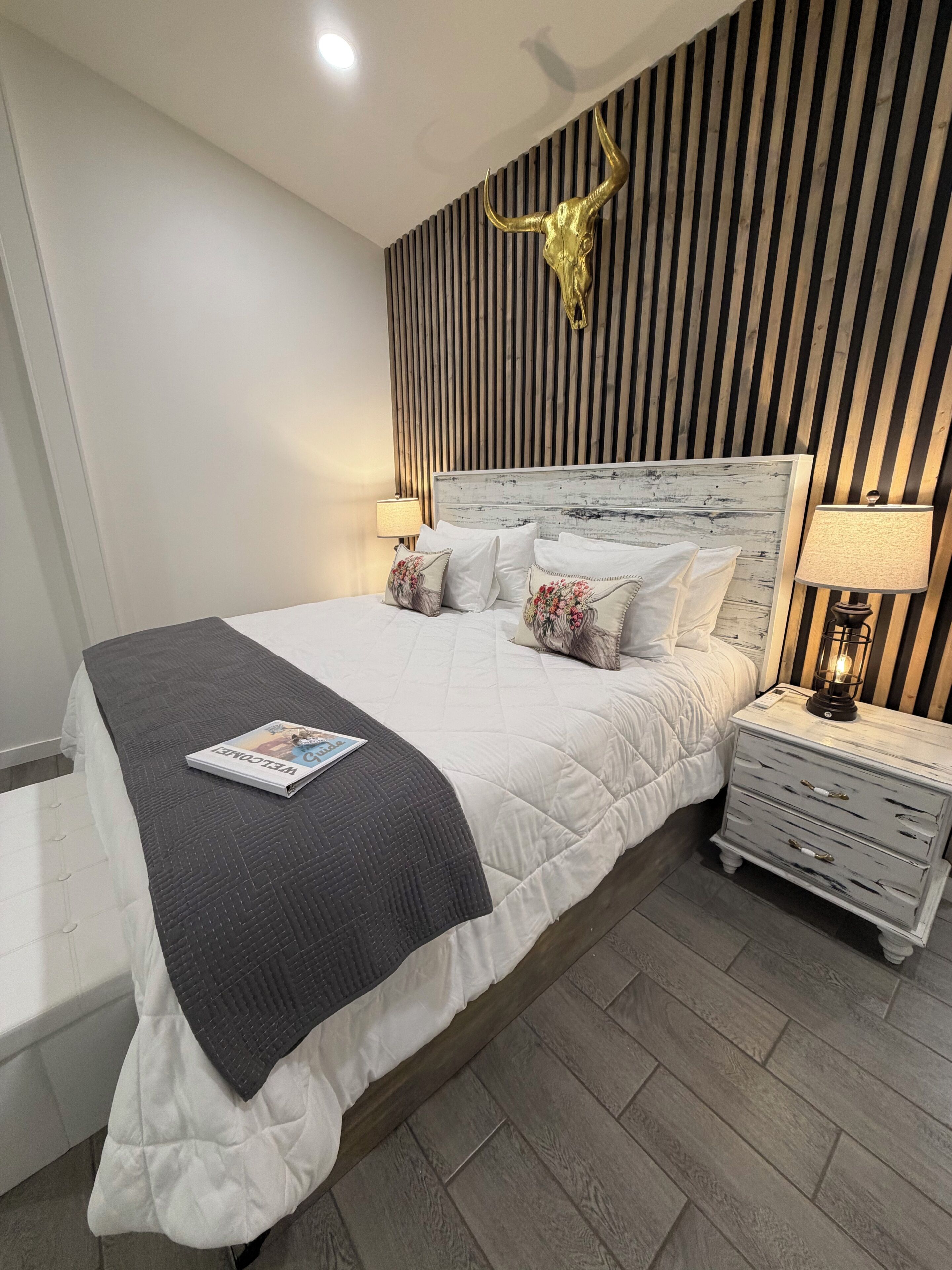Un dormitorio con cama, mesitas de noche, lámpara, una revista y una calavera decorativa en la pared.