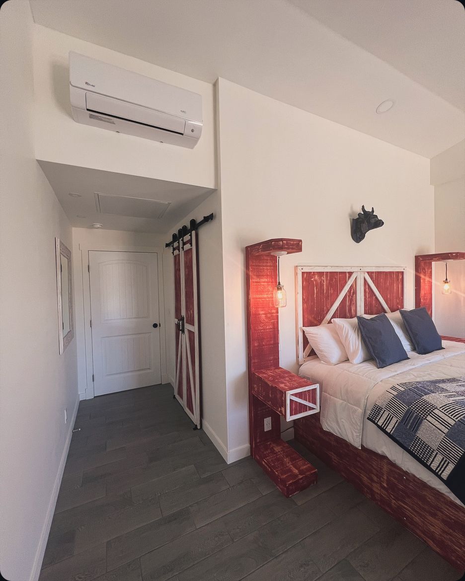 Un dormitorio con cama, aire acondicionado instalado en la pared, cabecero de madera, lámpara de pared y una puerta de estilo rústico.