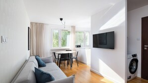 A modern living room with a grey sofa, a dining table with chairs, a washing machine, and large windows. - Metro Stokłosy 2-Bedroom Apartment (Warszawa)