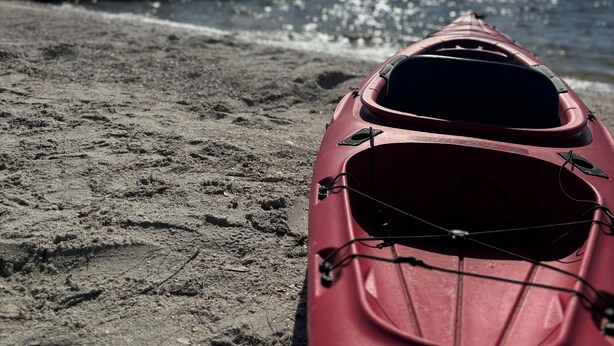 Un kayak rouge sur une plage de sable, avec une jetée en bois en arrière-plan et un ciel dégagé.