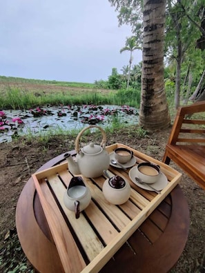 Een ronde houten tafel met een dienblad waarop een theepot, kopjes en een kom staan, buiten opstelling met een vijver en lotusbloemen op de achtergrond.
