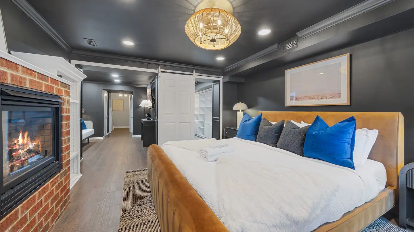 A modern bedroom with a fireplace, a large bed with blue and gray pillows, a wooden headboard, and a chandelier.