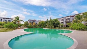 A large, curved swimming pool surrounded by a paved area with palm trees and buildings in the background. - Luxy Park Hotel & Apartments - MTown (Phu Quoc Island)