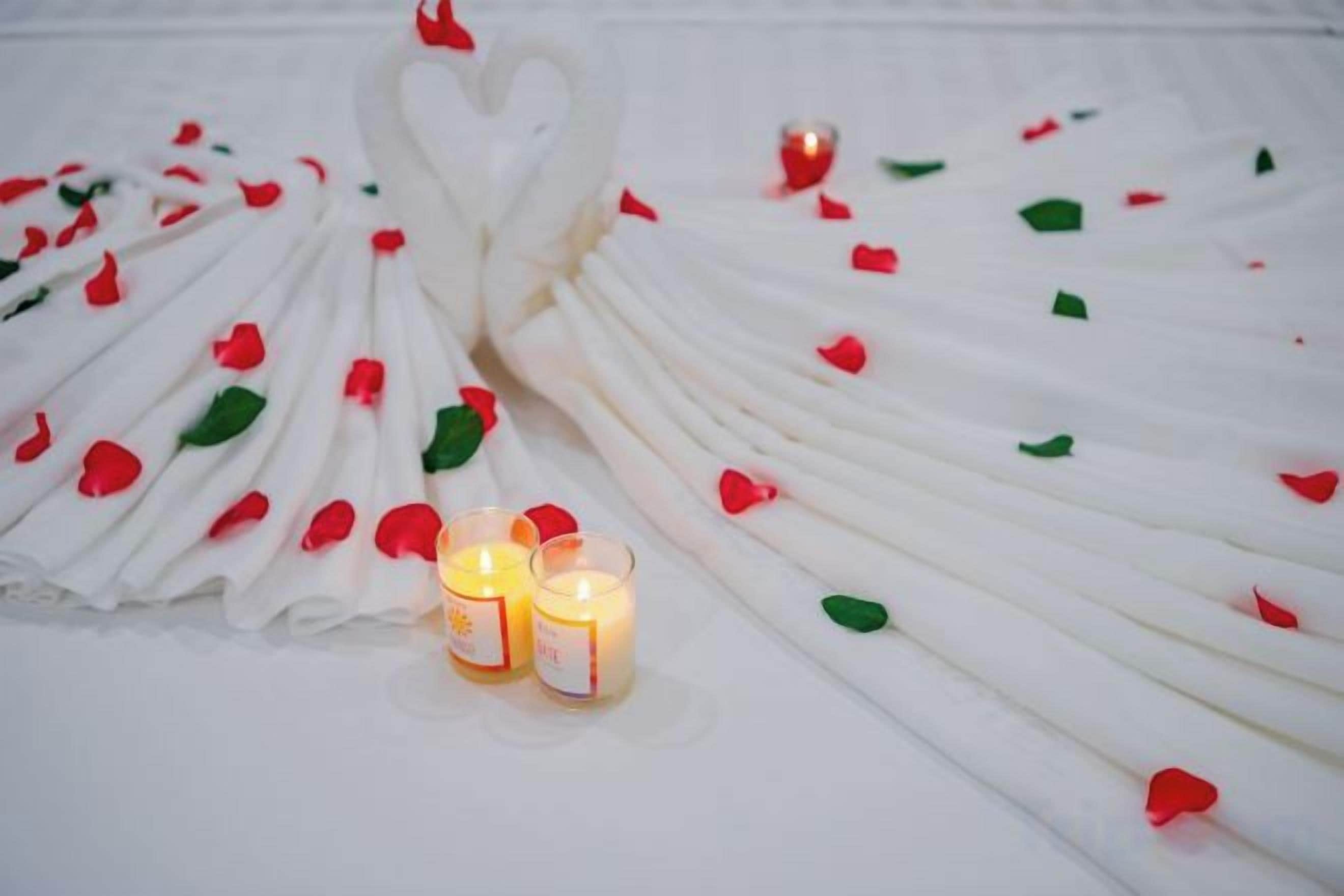 A white table with a heart-shaped arrangement of napkins and scattered rose petals, accompanied by two lit candles.