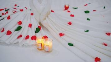 A white table with a heart-shaped arrangement of napkins and scattered rose petals, accompanied by two lit candles.
