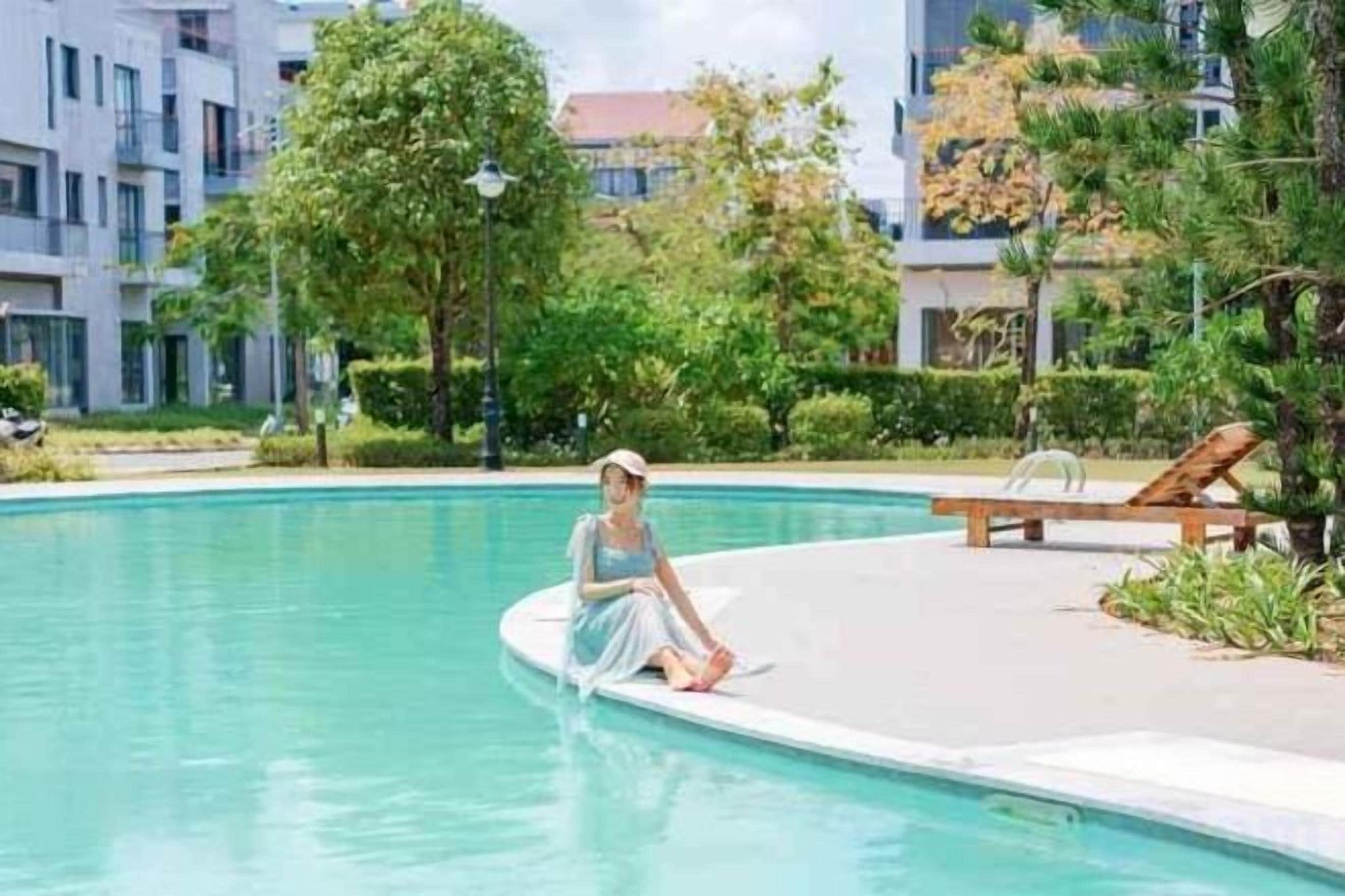 Uma pessoa sentada à beira da piscina com uma espreguiçadeira e um edifício ao fundo.