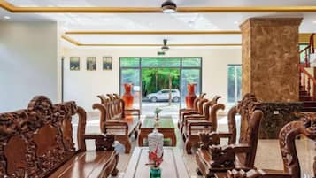 A lobby with ornate wooden furniture, a central table with a vase, and a view of the outdoors through large windows.