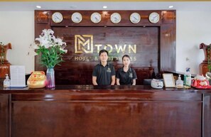 A reception desk with a wooden backdrop displaying the hotel name, clocks, and a vase of flowers. - Luxy Park Hotel & Apartments - MTown (Phu Quoc Island)